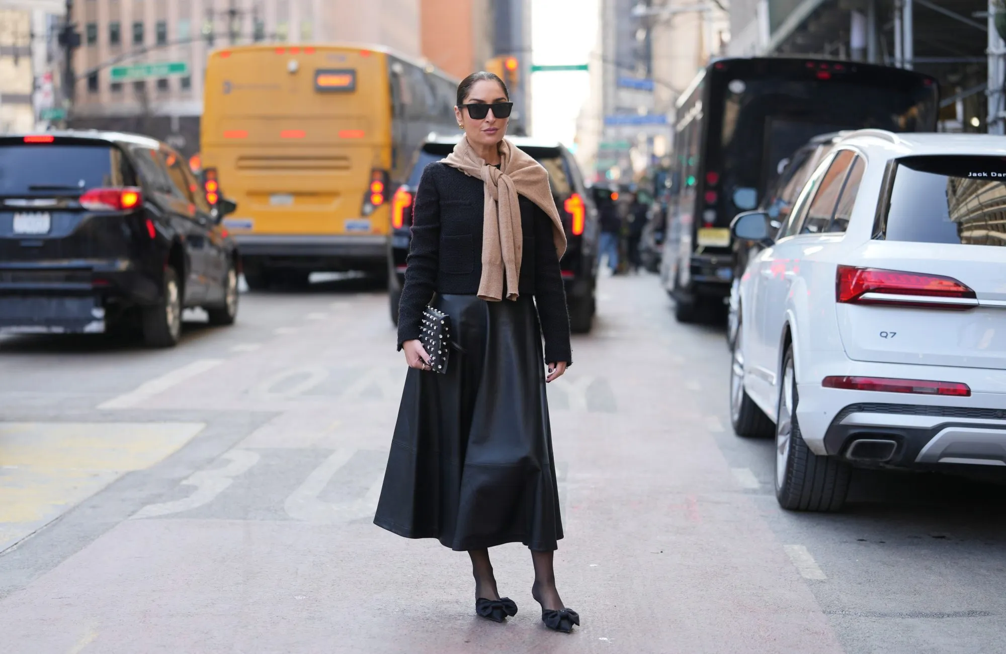 NEW YORK, NEW YORK - FEBRUARY 14: Geraldine Boublil wears a bun, black rectangular sunglasses, earrings, a black leather nailed / studded clutch bag, a beige knit sweater, a black textured jacket, a black leather midi skirt, black sheer tights, black pointed-toe flat shoes with a large bow toe cap, outside Altuzarra, during New York Fashion Week, on February 14, 2026 in New York City (Photo by Edward Berthelot/Getty Images)