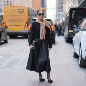 NEW YORK, NEW YORK - FEBRUARY 14: Geraldine Boublil wears a bun, black rectangular sunglasses, earrings, a black leather nailed / studded clutch bag, a beige knit sweater, a black textured jacket, a black leather midi skirt, black sheer tights, black pointed-toe flat shoes with a large bow toe cap, outside Altuzarra, during New York Fashion Week, on February 14, 2026 in New York City (Photo by Edward Berthelot/Getty Images)