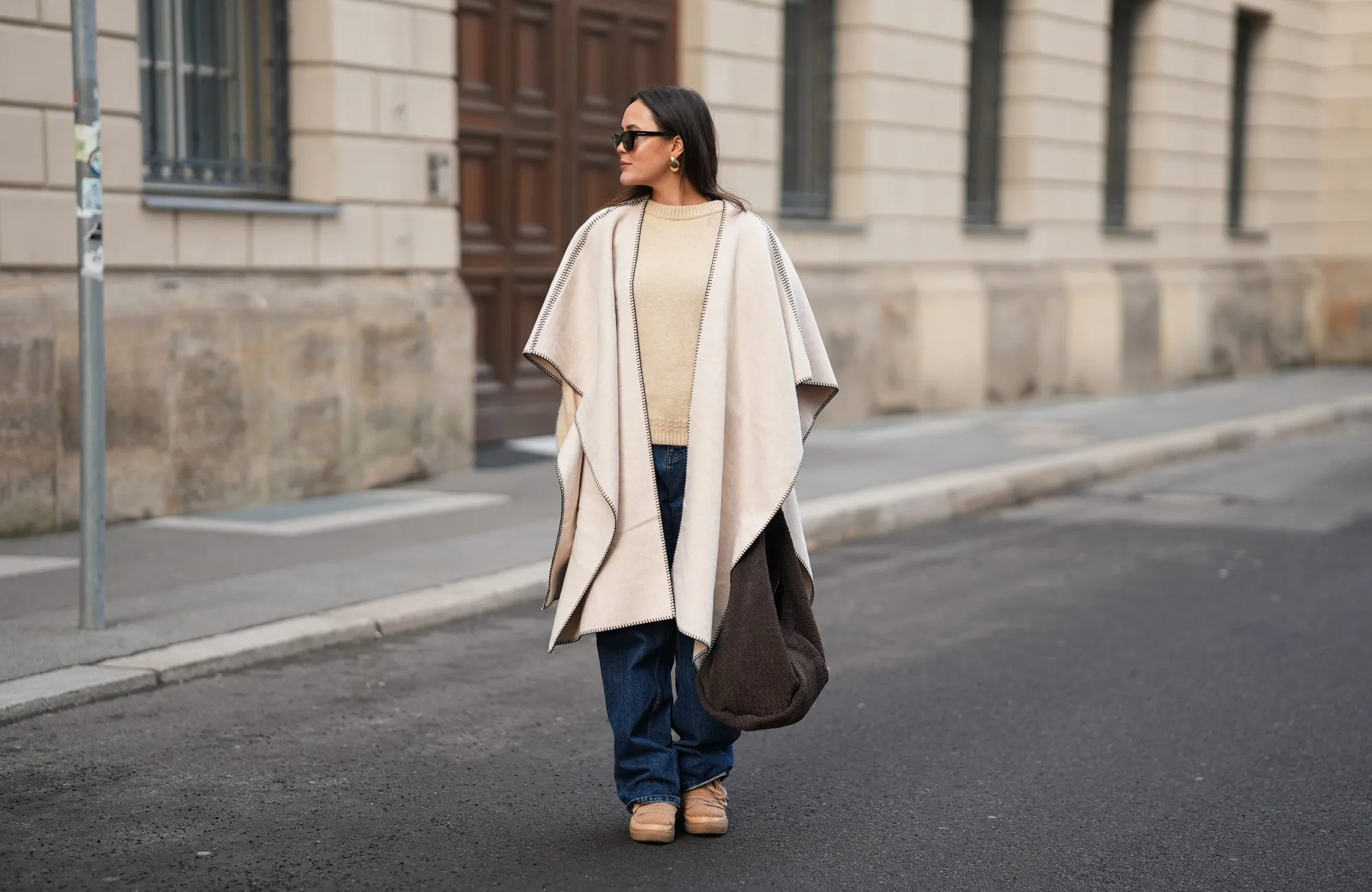 BERLIN, GERMANY - NOVEMBER 25: Laura Noltemeyer is seen wearing a cream oversized wool poncho with contrast whipstitch edging, a wide draped silhouette and open-front drape from Zara; layered underneath is a soft beige wool-blend knit sweater with a ribbed crew neckline and ribbed trims from Sézane; straight-leg dark-blue denim trousers with a classic fit from By Aylin Koenig; beige suede winter boots with a rounded toe, rope laces, tonal stitching and a thick rubber sole from Inuikii; a large textured dark-brown shoulder bag in a soft sculptural, slouchy shape and woven leather from Les Visionnaires x Michi from the Want; chunky sculptural gold earrings from Zara; black angular acetate sunglasses with a thick frame from Yves Saint Laurent; her dark brown hair is worn straight with a center part, tucked loosely behind the ears on November 25, 2025 in Berlin, Germany. (Photo by Moritz Scholz/Getty Images)