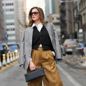 NEW YORK, NEW YORK - FEBRUARY 14: A guest wears loose hair, black sunglasses, gold earrings, rings, a black leather Kelly clutch bag from Hermes, a gray plaid blazer jacket, a black knit V-neck cardigan with gold buttons, a white collared shirt, brown wide-leg pants, a gold chain belt, a white pearls belt, outside Altuzarra, during New York Fashion Week, on February 14, 2026 in New York City (Photo by Edward Berthelot/Getty Images)