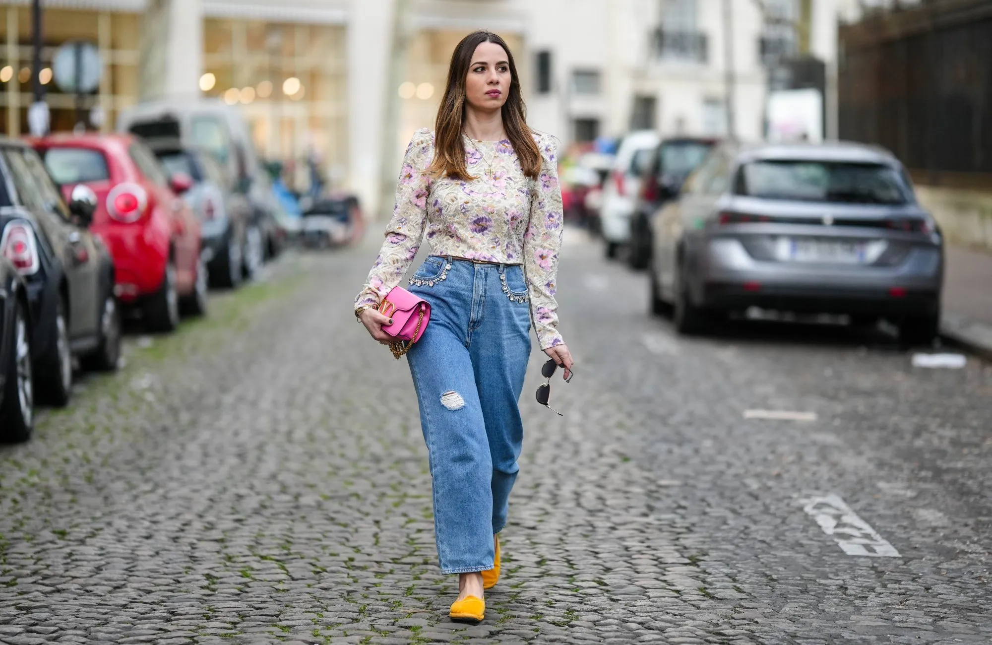 PARIS, FRANCE - MARCH 25: Maria Rosaria Rizzo wears a gold long chain with large cross pendant necklace, a beige with purple and pink tie and dye print pattern long sleeves / puffy shoulder blouse, a neon pink shiny leather long clutch with gold large logo buckle from Valentino, a brown beige shiny leather with gold mini logo buckle belt from Valentino, blue faded denim ripper large pants, yellow gold velvet loafers , during a street style fashion photo session, on March 25, 2023 in Paris, France. (Photo by Edward Berthelot/Getty Images)