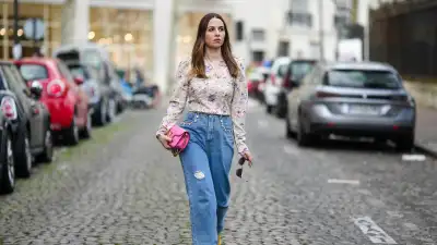 PARIS, FRANCE - MARCH 25: Maria Rosaria Rizzo wears a gold long chain with large cross pendant necklace, a beige with purple and pink tie and dye print pattern long sleeves / puffy shoulder blouse, a neon pink shiny leather long clutch with gold large logo buckle from Valentino, a brown beige shiny leather with gold mini logo buckle belt from Valentino, blue faded denim ripper large pants, yellow gold velvet loafers , during a street style fashion photo session, on March 25, 2023 in Paris, France. (Photo by Edward Berthelot/Getty Images)