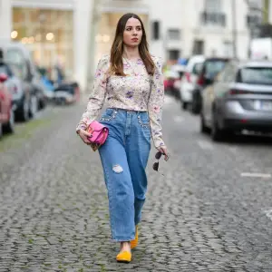 PARIS, FRANCE - MARCH 25: Maria Rosaria Rizzo wears a gold long chain with large cross pendant necklace, a beige with purple and pink tie and dye print pattern long sleeves / puffy shoulder blouse, a neon pink shiny leather long clutch with gold large logo buckle from Valentino, a brown beige shiny leather with gold mini logo buckle belt from Valentino, blue faded denim ripper large pants, yellow gold velvet loafers , during a street style fashion photo session, on March 25, 2023 in Paris, France. (Photo by Edward Berthelot/Getty Images)