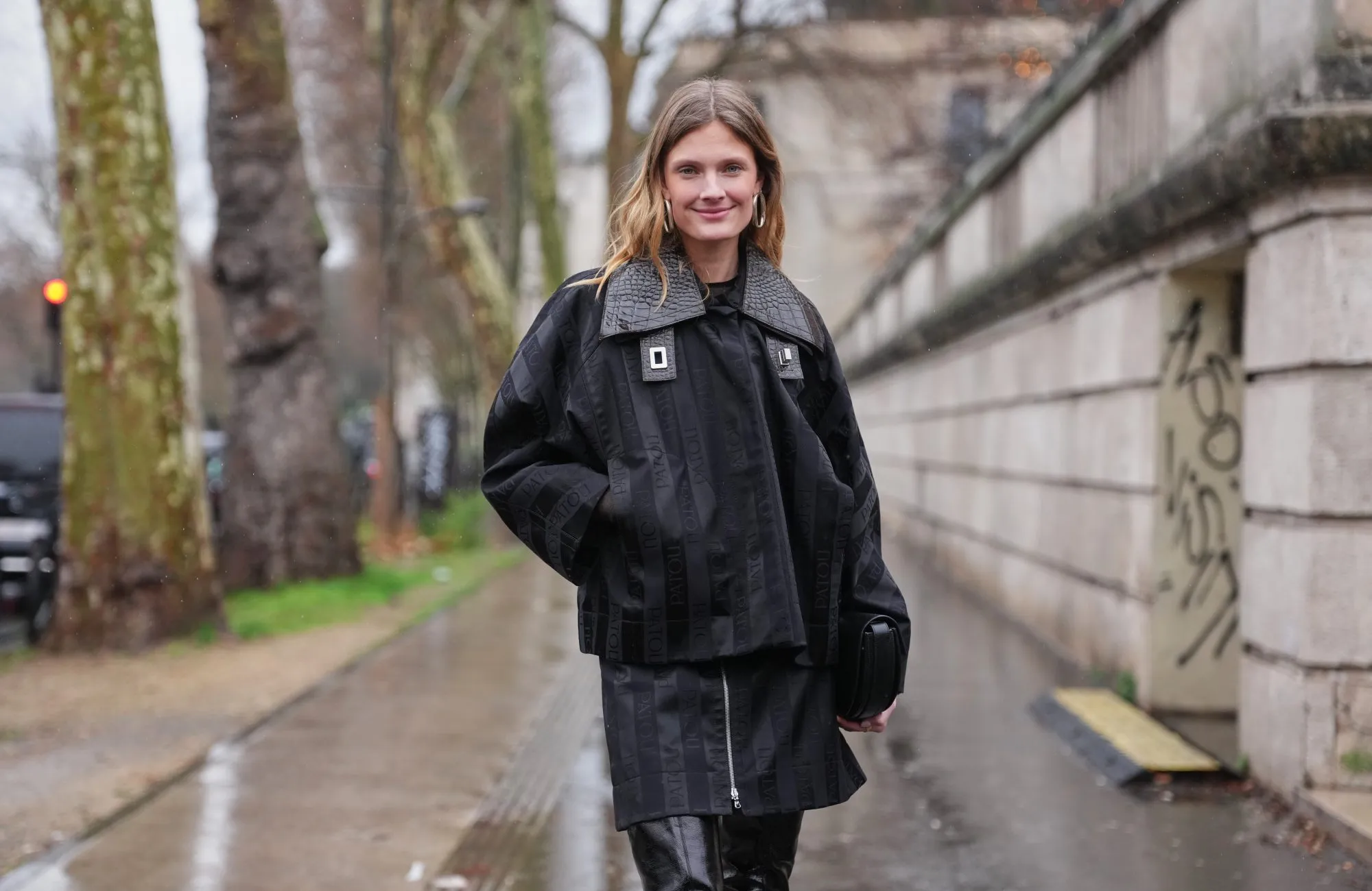 PARIS, FRANCE - JANUARY 25: Constance Jablonski wears long wavy blonde hair with a center part, metal hoop earrings, a handheld black leather clutch bag, a black oversized jacket with tonal PATOU logo-jacquard striping, textured leather collar and metal buckle details layered over a black top, a matching mini skirt, black glossy leather skinny boot, outside Patou, during Paris Fashion Week - Menswear Fall/Winter 2026-2027, on January 25, 2026 in Paris, France (Photo by Edward Berthelot/Getty Images)