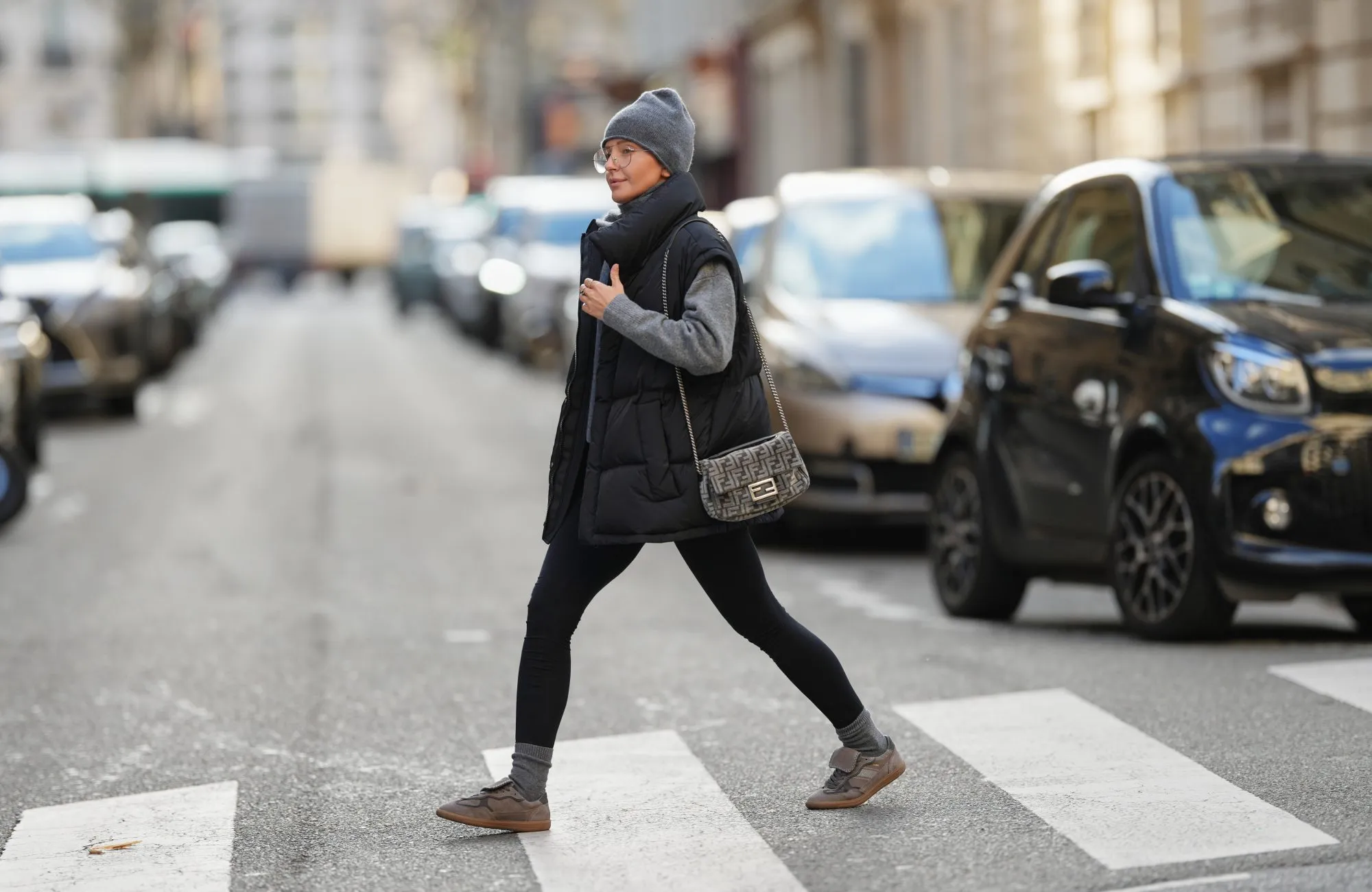PARIS, FRANCE - JANUARY 15: Diane Batoukina wears long straight brown hair tucked under a ribbed grey cashmere beanie hat by Mango, Kaleos rectangular eyeglasses with thin dark frames, a matching grey cashmere scarf by Mango wrapped around the neck, a medium grey knit sweater by Zara with a high neckline, a black quilted padded sleeveless puffer jacket by WRSTBHVR featuring a matte finish and snap-button front, black stretch leggings by Zara with a smooth matte texture, ribbed grey cashmere socks by Mango worn above the ankle, grey and taupe lace-up sneakers by Alohas with a low-profile rubber sole, and a small grey Fendi monogram shoulder bag with a flap closure and metal chain strap carried at the side, during a street style fashion photo session, on January 15, 2026 in Paris, France. (Photo by Edward Berthelot/Getty Images)