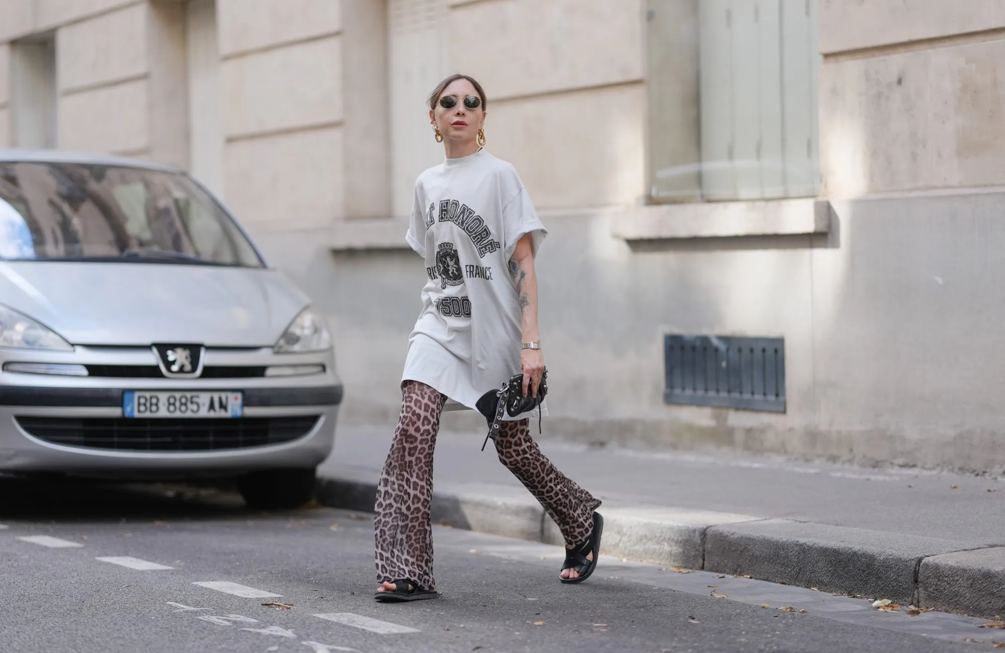 PARIS, FRANCE - JULY 17: Emy Venturini wears an oversized white graphic T-shirt dress by Balenciaga paired with flared leopard print trousers by Calzedonia. Black open-toe flat sandals by Prada complete the look. Accessories include a black leather clutch by Balenciaga, a silver Cartier wristwatch, gold sculptural earrings by Rabanne, and round sunglasses by Ray-Ban. Hair is blond and styled in a low ponytail with a center part, and lips are painted with lipstick in a bold red shade, during a street style fashion photo session, on July 17, 2025 in Paris, France. (Photo by Edward Berthelot/Getty Images)