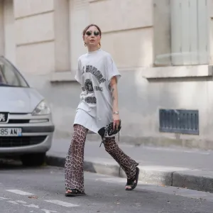 PARIS, FRANCE - JULY 17: Emy Venturini wears an oversized white graphic T-shirt dress by Balenciaga paired with flared leopard print trousers by Calzedonia. Black open-toe flat sandals by Prada complete the look. Accessories include a black leather clutch by Balenciaga, a silver Cartier wristwatch, gold sculptural earrings by Rabanne, and round sunglasses by Ray-Ban. Hair is blond and styled in a low ponytail with a center part, and lips are painted with lipstick in a bold red shade, during a street style fashion photo session, on July 17, 2025 in Paris, France. (Photo by Edward Berthelot/Getty Images)