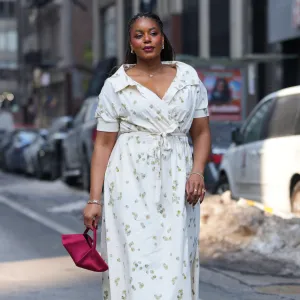 NEW YORK, NEW YORK - FEBRUARY 14: A guest wears braided hair, earrings, necklace, gold rings, a pink satin clutch bag, a white V-neck wrap dress with a tie waist and a floral print, gold metallic open toe sandals shoes, outside Altuzarra, during New York Fashion Week, on February 14, 2026 in New York City (Photo by Edward Berthelot/Getty Images)
