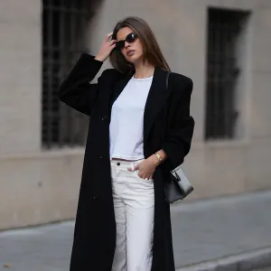 MUNICH, GERMANY - NOVEMBER 5: Iva Nikolina is seen wearing a long black wool coat with structured shoulders and a straight silhouette by Almada Label; a white fitted cotton crewneck T-shirt by Re/Done; white wide-leg jeans in a straight leg cut by The Row; black polished leather loafers with a gold logo detail by Saint Laurent; a structured black leather “Kelly” handbag with silver hardware by Hermès; a small gold „Panthére“ watch by Cartier; narrow oval black sunglasses by Selima Optique; her brown hair is worn straight and parted in the middle on November 5, 2025 in Munich, Germany. (Photo by Moritz Scholz/Getty Images)