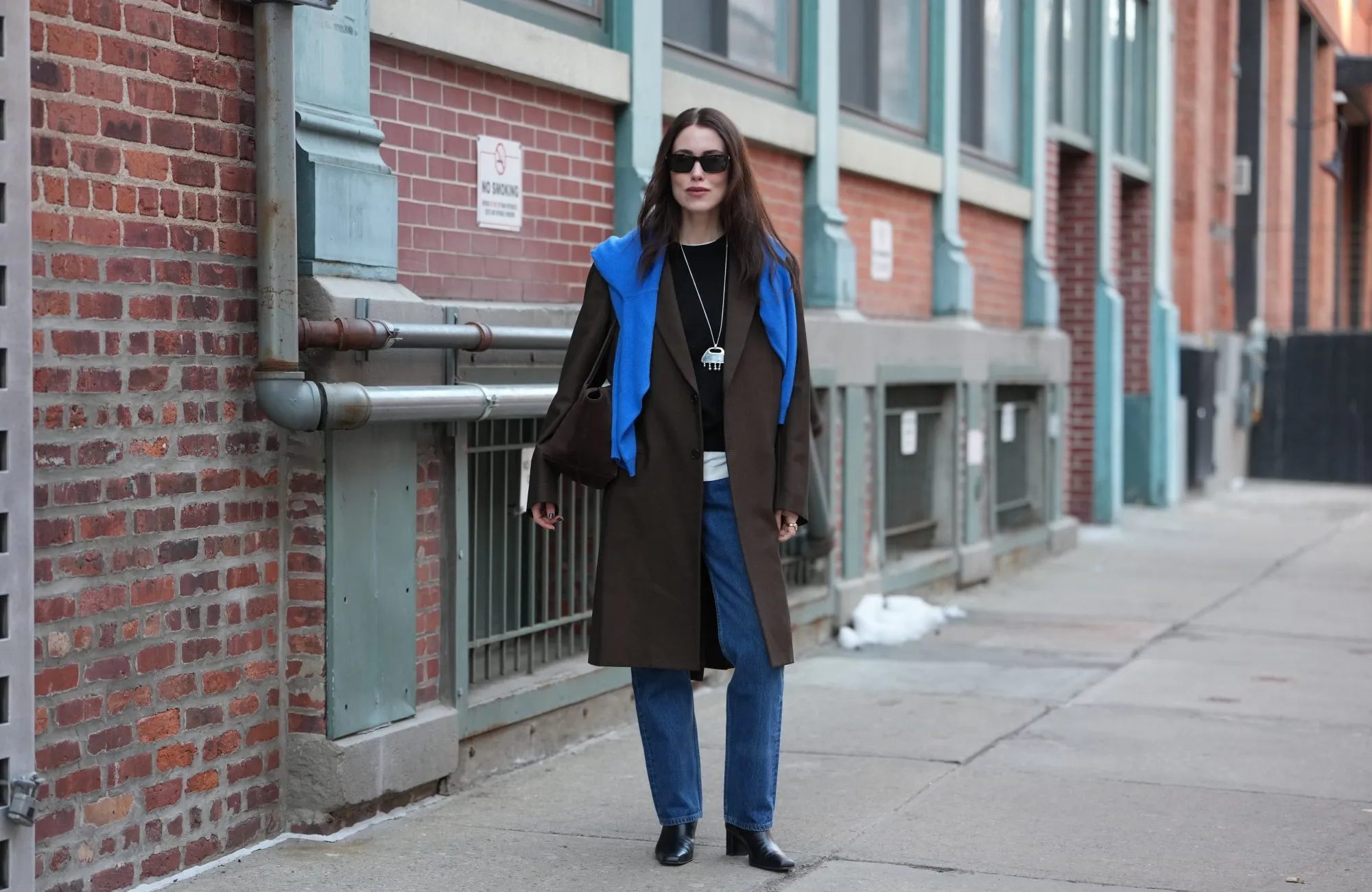 NEW YORK, NEW YORK - FEBRUARY 13: Annabel Rosendahl wears long dark brown hair, black rectangular sunglasses, a long silver pendant necklace with pink print pattern pendant, a dark brown wool coat, a blue wool pullover, a black crew neck sweater, a white shirt, blue denim straight-leg jeans pants, black leather block heels / ankle boots shoes, outside Ulla Johnson, during New York Fashion Week, on February 13, 2026 in New York City (Photo by Edward Berthelot/Getty Images)