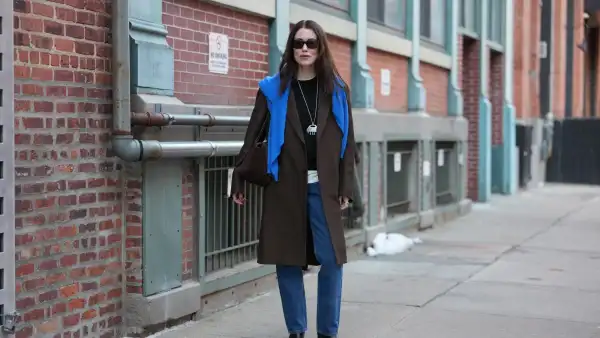 NEW YORK, NEW YORK - FEBRUARY 13: Annabel Rosendahl wears long dark brown hair, black rectangular sunglasses, a long silver pendant necklace with pink print pattern pendant, a dark brown wool coat, a blue wool pullover, a black crew neck sweater, a white shirt, blue denim straight-leg jeans pants, black leather block heels / ankle boots shoes, outside Ulla Johnson, during New York Fashion Week, on February 13, 2026 in New York City (Photo by Edward Berthelot/Getty Images)
