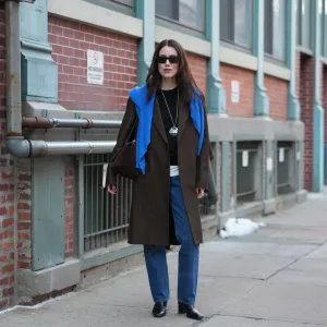 NEW YORK, NEW YORK - FEBRUARY 13: Annabel Rosendahl wears long dark brown hair, black rectangular sunglasses, a long silver pendant necklace with pink print pattern pendant, a dark brown wool coat, a blue wool pullover, a black crew neck sweater, a white shirt, blue denim straight-leg jeans pants, black leather block heels / ankle boots shoes, outside Ulla Johnson, during New York Fashion Week, on February 13, 2026 in New York City (Photo by Edward Berthelot/Getty Images)