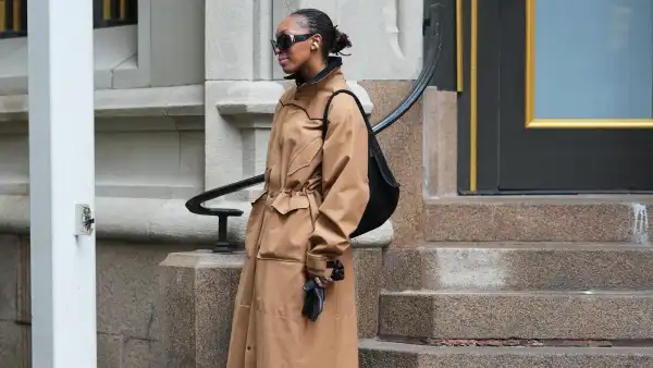 NEW YORK, NEW YORK - FEBRUARY 15: A guest wears black hair in a bun, black wraparound sunglasses, black wireless earbuds, a black woven backpack bag, a tan cotton high neck trench coat, black leather gloves, outside Diotima, during New York Fashion Week, on February 15, 2026 in New York City (Photo by Edward Berthelot/Getty Images)