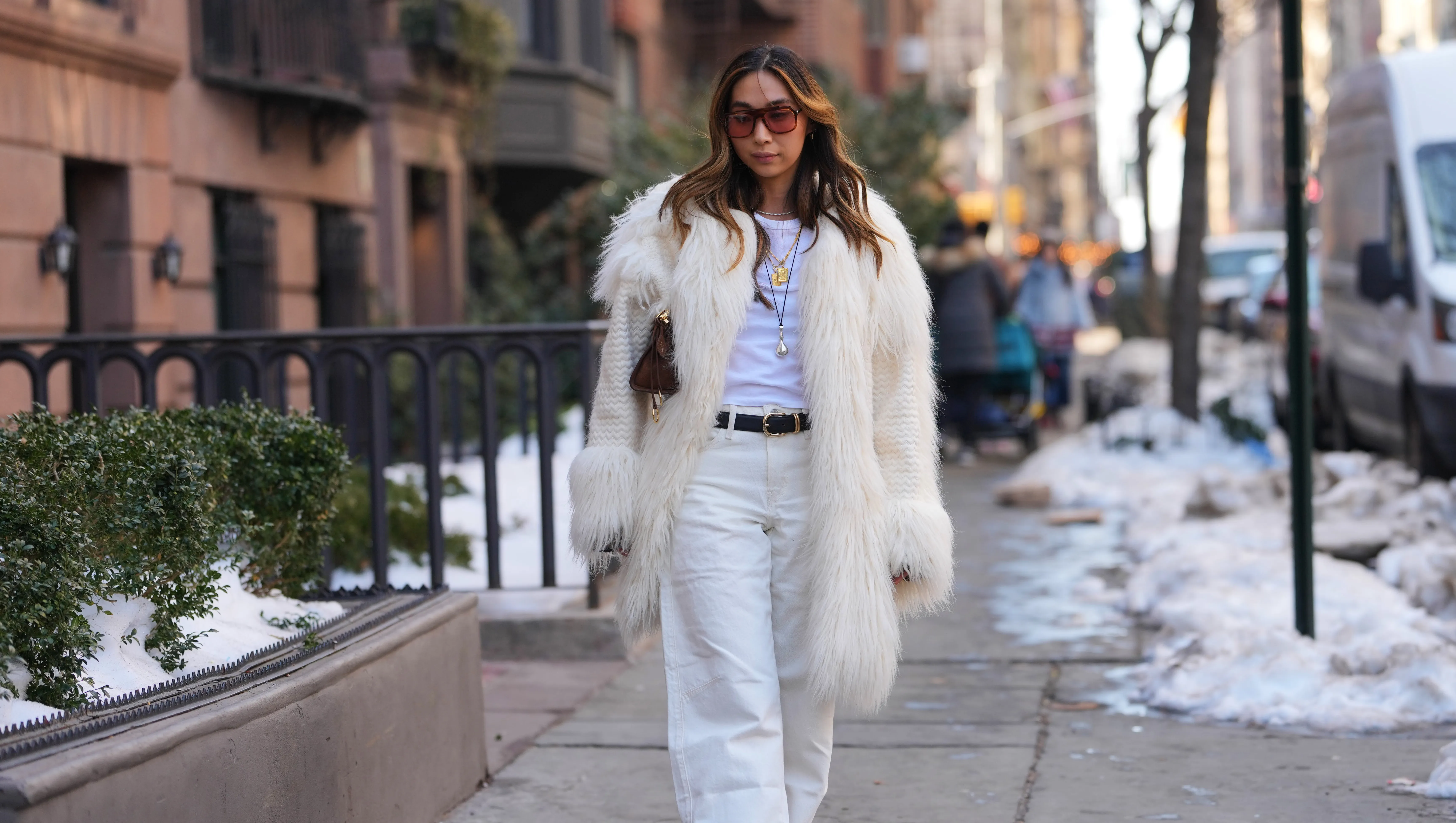 NEW YORK, NEW YORK - FEBRUARY 14: A guest wears long wavy brown hair, brown tinted sunglasses, gold pendant necklaces, a brown leather mini bag from Alaia, a white braided fabric and faux fur coat, a white crew neck t-shirt, white straight-leg jeans pants, a black leather belt, brown suede pointed-toe ankle boots shoes, outside Anna Sui, during New York Fashion Week, on February, 14 2026 in New York City (Photo by Edward Berthelot/Getty Images)