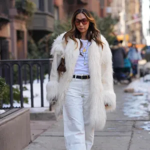 NEW YORK, NEW YORK - FEBRUARY 14: A guest wears long wavy brown hair, brown tinted sunglasses, gold pendant necklaces, a brown leather mini bag from Alaia, a white braided fabric and faux fur coat, a white crew neck t-shirt, white straight-leg jeans pants, a black leather belt, brown suede pointed-toe ankle boots shoes, outside Anna Sui, during New York Fashion Week, on February, 14 2026 in New York City (Photo by Edward Berthelot/Getty Images)