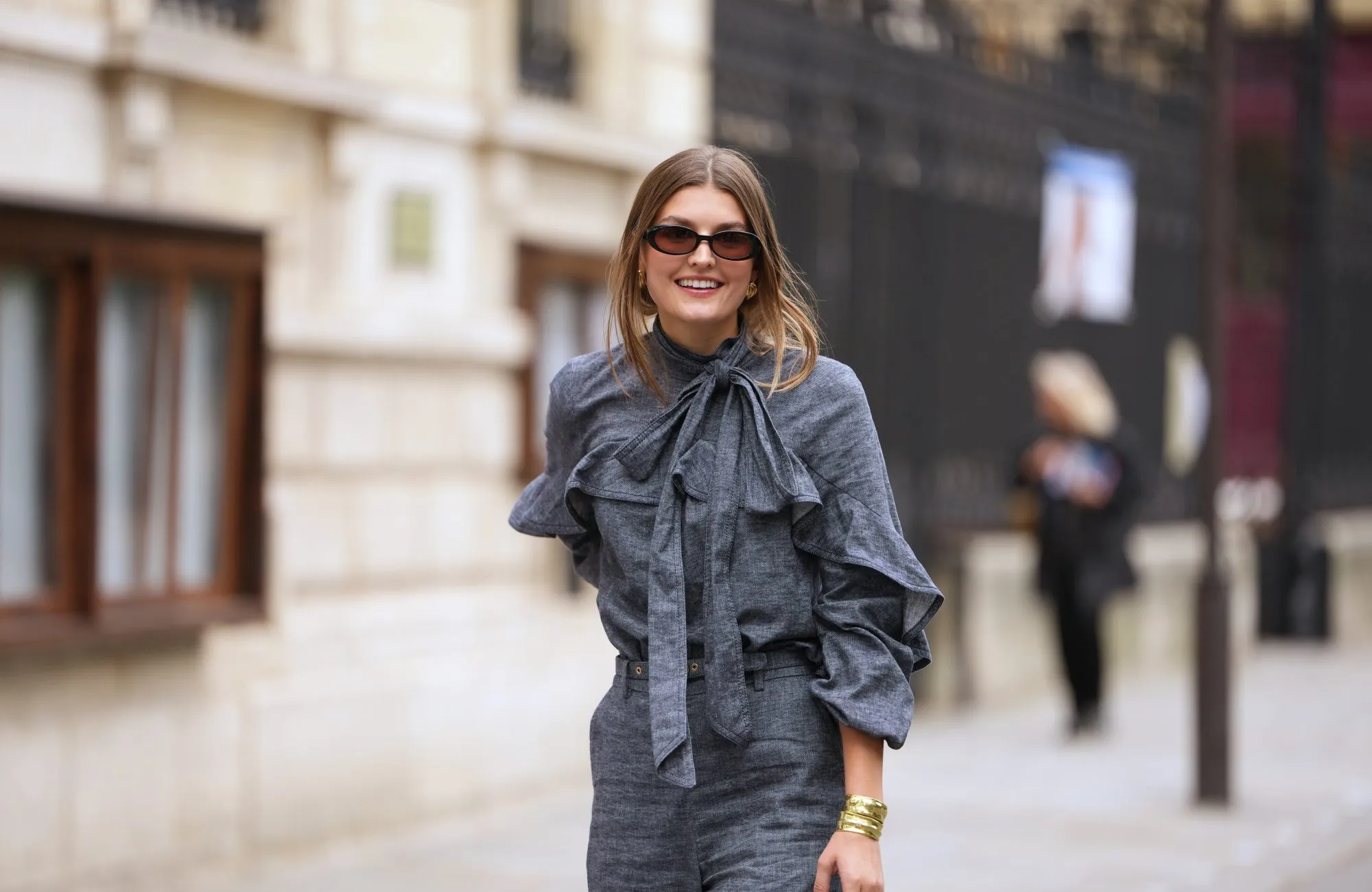 PARIS, FRANCE - OCTOBER 06: A guest wears black square sunglasses, a gray blue denim ruffled high neck / knot bow tie / long sleeves blouse, matching gray blue flared suit pants, outside Zimmermann, during Paris Fashion Week - Womenswear Spring Summer 2026, on October 06, 2025 in Paris, France (Photo by Edward Berthelot/Getty Images)