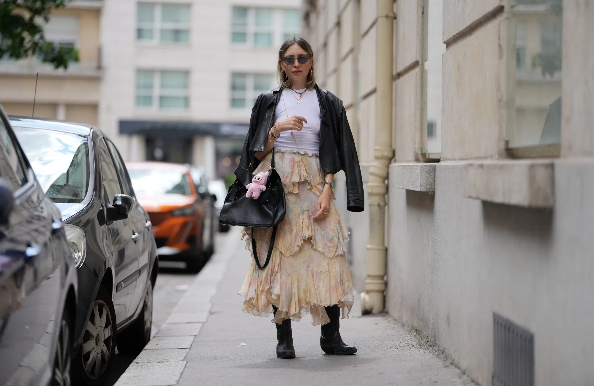 PARIS, FRANCE - JUNE 06: Emy Venturini wears a black leather jacket draped over the shoulders, paired with a white ribbed top. The skirt is a layered, ruffled design in a pastel floral pattern with shades of pink, yellow, and green. Accessories include small hoop earrings and a necklace with a pendant. Sunglasses with dark lenses are worn. The black leather handbag features a plush pink Labubu fluffy monster toy attached. Footwear consists of black cowboy boots, during a street style fashion photo session, on June 06, 2025 in Paris, France (Photo by Edward Berthelot/Getty Images)