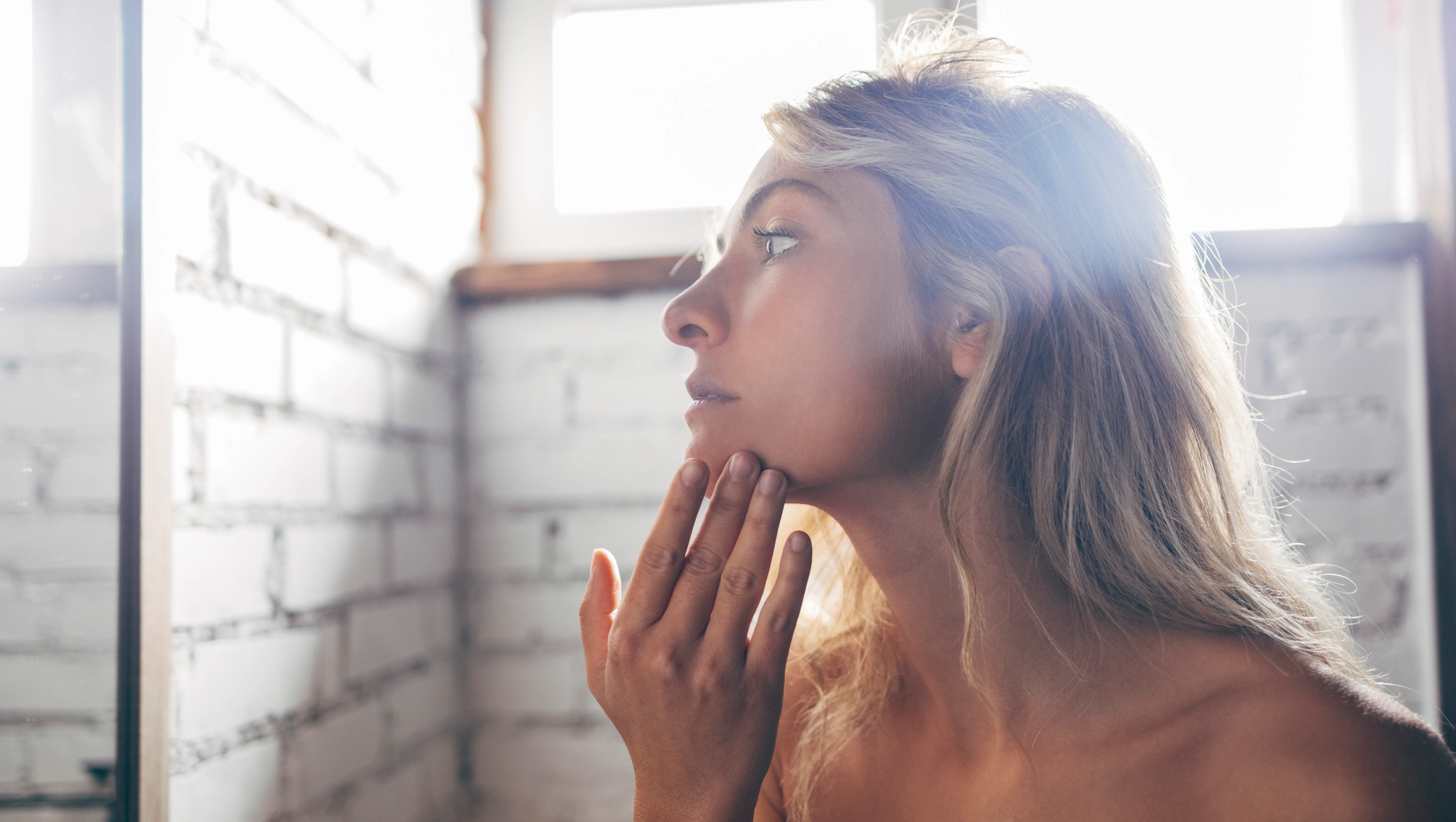 A portrait of a young Caucasian woman standing in the bathroom wrapped in a gray towel and applying face cream.