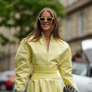 COPENHAGEN, DENMARK - AUGUST 04: Nina Sandbech wears a light yellow dress with a structured, high neckline and long sleeves. The dress features a fitted waist and midi-length skirt accentuated by a large yellow leather belt and a flared. Sunglasses with a matching yellow frame are worn. A black and white patterned clutch is held in one hand. The outfit is accessorized with gold bracelets on the wrist. Black and white zebra-patterned heels complete the ensemble, outside OperaSport, during Copenhagen Fashion Week, on August 04, 2025 in Copenhagen, Denmark (Photo by Edward Berthelot/Getty Images)