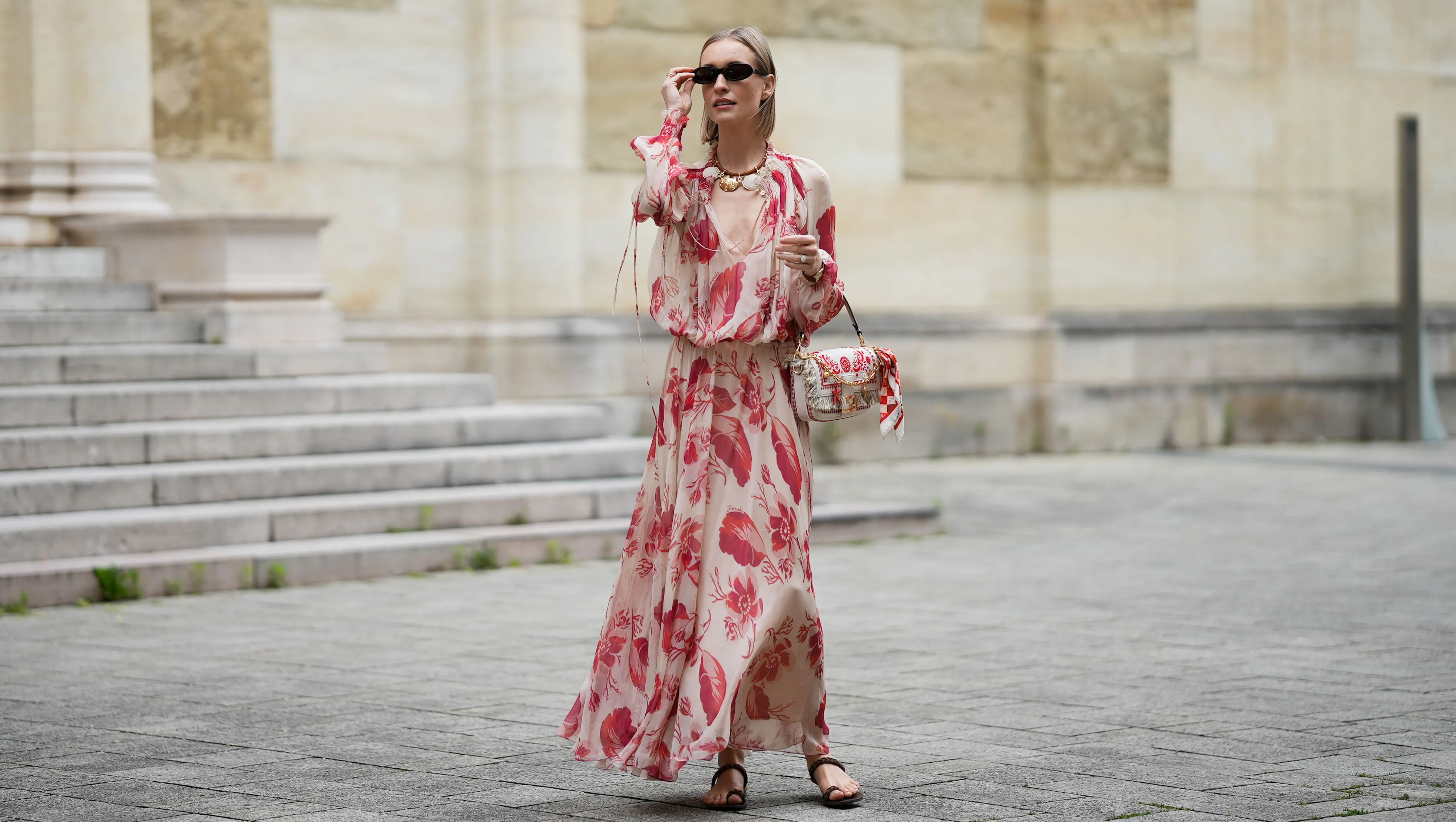 MUNICH, GERMANY - MAY 29: Marlies Pia Pfeifhofer is seen wearing a light beige silk chiffon long sleeve maxi dress with red and pink prints of feathers and flowers, drawstring waist, deep V-neck with front tie and smocking details on the collar and cuffs from FENDI, a natural-colored Baguette canvas bag with coral bandana embroidery, small fringes on the flap, calfskin leather straps, golden hardware and logo detail as well as an additional golden chain metal charm with marine-inspired pendants and a red, pink and white bandana charm with golden logo clip from FENDI, a pair of chocolate brown leather gladiator sandals with a braided leather toe strap and one main strap from Alex Rivière x Manebi, a Louis Tortoise Brown sunglasses with slim and oval shape from Corlin, a gold-colored metal and brown nappa leather Forever Fendi necklace with shell-shaped pendants from FENDI, a brown leather Tank Louis watch with yellow gold rectangular watch case from Cartier, a silver diamond ring on May 29, 2025 in Munich, Germany. (Photo by Moritz Scholz/Getty Images)