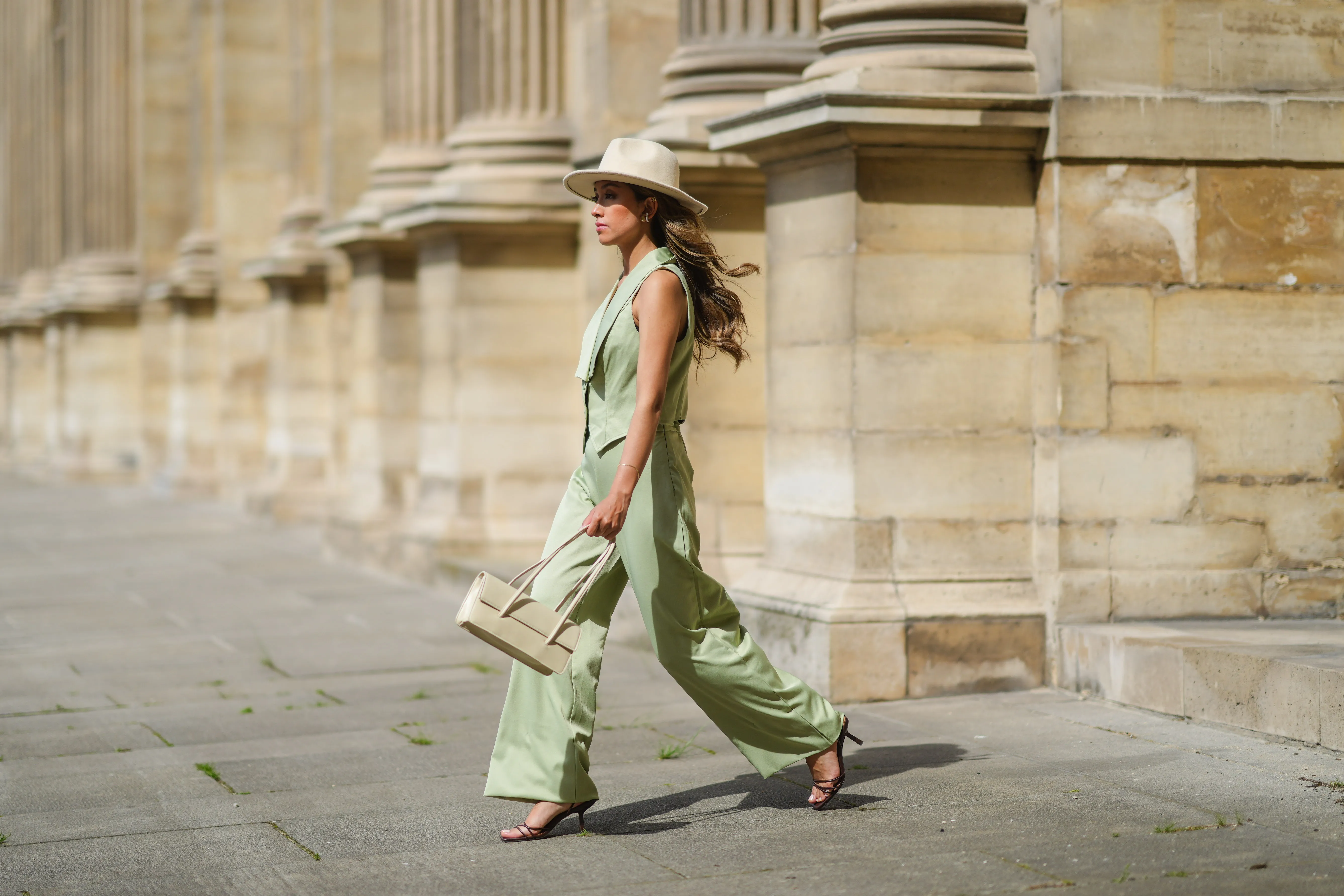 Angela Gonzalez wears a beige suede Lack Of Color hat, earrings, a black off shoulder top, a pale green buttoned blazer jacket sleeveless from Nasty Gal, pale green matching Nasty Gal flared pants, a gold chain bracelet, a beige shiny leather Mango handbag, brown shiny leather strappy hells Na-Kd sandals, on May 29, 2021 in Paris, France.
