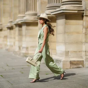 Angela Gonzalez wears a beige suede Lack Of Color hat, earrings, a black off shoulder top, a pale green buttoned blazer jacket sleeveless from Nasty Gal, pale green matching Nasty Gal flared pants, a gold chain bracelet, a beige shiny leather Mango handbag, brown shiny leather strappy hells Na-Kd sandals, on May 29, 2021 in Paris, France.