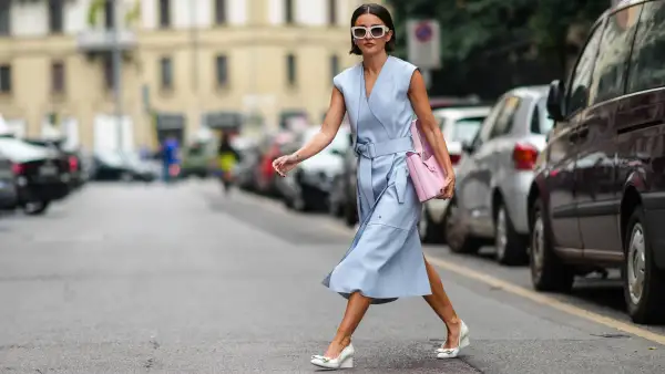 Alexandra Pereira wears white sunglasses, silver and rhinestones earrings, a pale blue shiny leather V-neck / sleeveless / belted / slit midi dress, a pale pink handbag from Salvatore Ferragamo, white leather block heels shoes with embroidered knot on the toe-cap, rings, outside the Ferragamo fashion show during the Milan Fashion Week - Spring / Summer 2022 on September 25, 2021 in Milan, Italy.