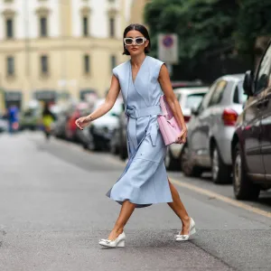 Alexandra Pereira wears white sunglasses, silver and rhinestones earrings, a pale blue shiny leather V-neck / sleeveless / belted / slit midi dress, a pale pink handbag from Salvatore Ferragamo, white leather block heels shoes with embroidered knot on the toe-cap, rings, outside the Ferragamo fashion show during the Milan Fashion Week - Spring / Summer 2022 on September 25, 2021 in Milan, Italy.