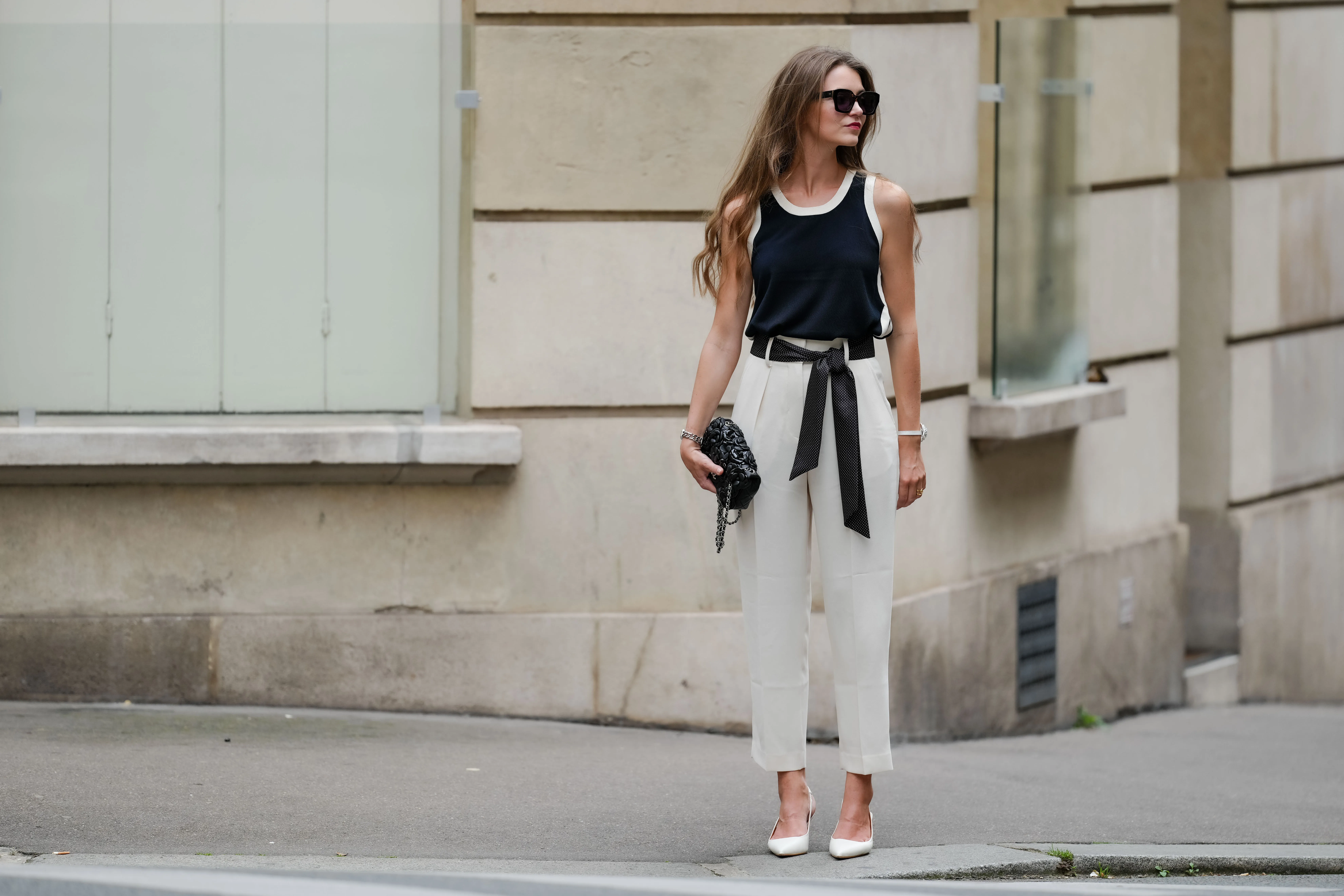 Segolene Hyppolite wears sunglasses, red lipstick / make-up, a dark navy blue / black and white tank top from Tara Jarmon, a black Chanel bag with floral textures / details, a Chanel watch, a black and white polka dots printed knot / scarf belt, a white floral shaped bracelet, white cropped pants from Tara Jarmon, white pumps / pointed shoes from Jonak, during a street style fashion photo session, on May 12, 2024 in Paris, France.