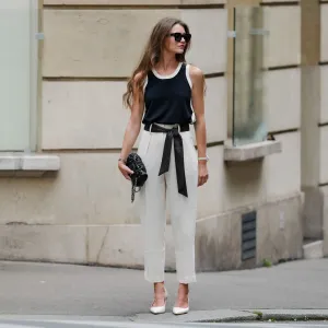 Segolene Hyppolite wears sunglasses, red lipstick / make-up, a dark navy blue / black and white tank top from Tara Jarmon, a black Chanel bag with floral textures / details, a Chanel watch, a black and white polka dots printed knot / scarf belt, a white floral shaped bracelet, white cropped pants from Tara Jarmon, white pumps / pointed shoes from Jonak, during a street style fashion photo session, on May 12, 2024 in Paris, France.
