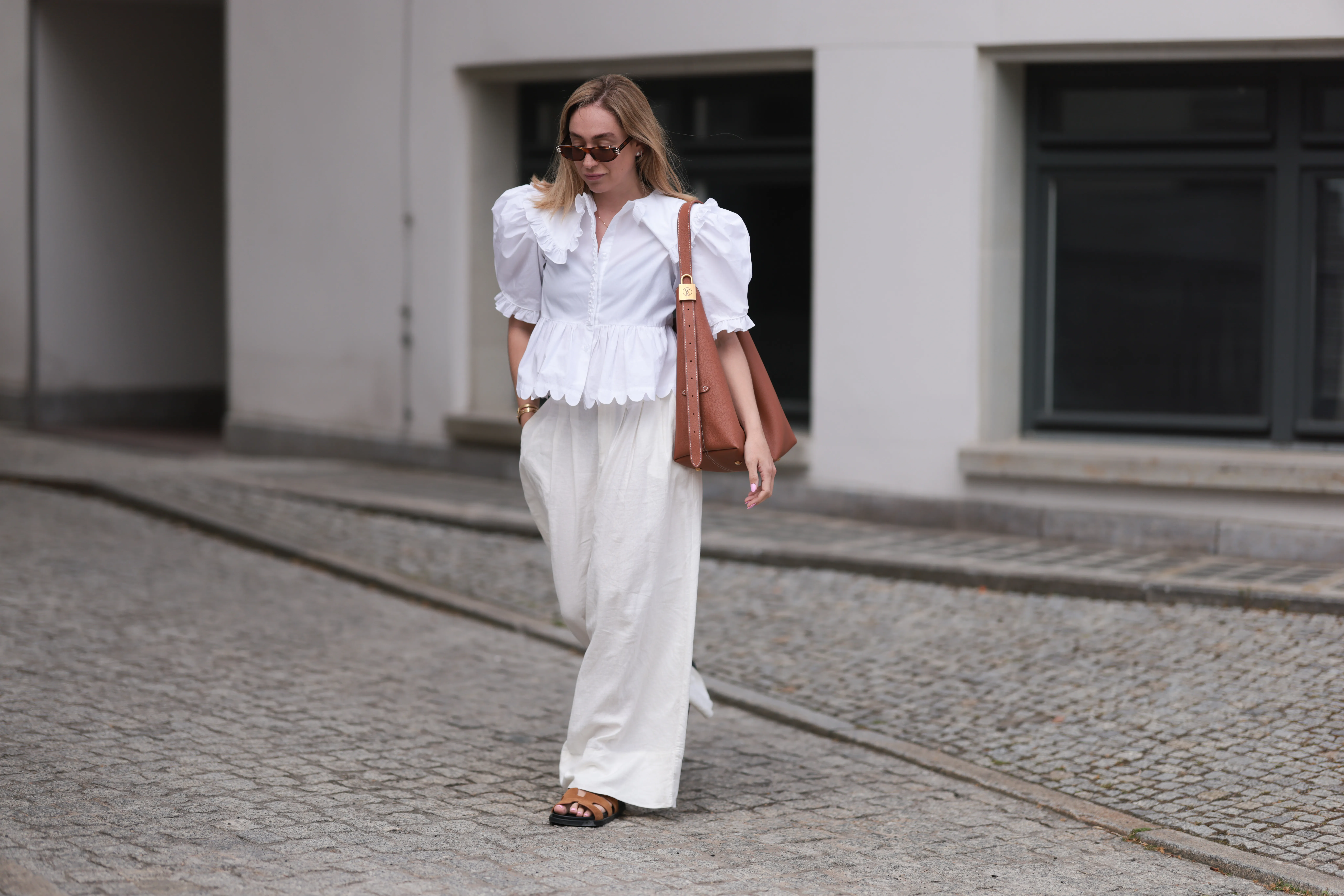 BERLIN, GERMANY - JUNE 11: Sonia Lyson is seen wearing sunglasses with havana frame from Givenchy, silver earrings, a golden necklace, a white blouse with puffed sleeves and ruffled details from Horror Vacui, cream-white wide linen pants from Zara, the “Low Key Hobo” bag in brown leather from Louis Vuitton and brown suede sandals from Hermès on June 11, 2024 in Berlin, Germany. (Photo by Jeremy Moeller/Getty Images)