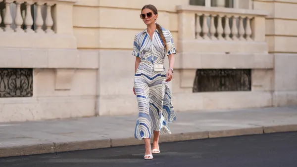 Diane Batoukina wears a white silk midi dress with blue and navy graphic stripe print, short sleeves, and a pointed collar by Sandro. A matching Sandro silk scarf is tied loosely around the neckline. A white mini flap bag with a chain strap and gold hardware by Chanel is worn crossbody. Footwear consists of white open-toe heeled sandals by Aeyde. Dior square sunglasses with dark gradient lenses complete the look. Long, straight brown hair is styled with a center part. Lips are subtly tinted and skin appears smooth and even. Full body shot, during a street style fashion photo session, on April 28, 2025 in Paris, France.