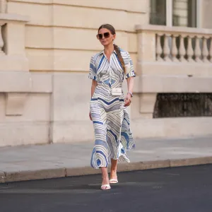 Diane Batoukina wears a white silk midi dress with blue and navy graphic stripe print, short sleeves, and a pointed collar by Sandro. A matching Sandro silk scarf is tied loosely around the neckline. A white mini flap bag with a chain strap and gold hardware by Chanel is worn crossbody. Footwear consists of white open-toe heeled sandals by Aeyde. Dior square sunglasses with dark gradient lenses complete the look. Long, straight brown hair is styled with a center part. Lips are subtly tinted and skin appears smooth and even. Full body shot, during a street style fashion photo session, on April 28, 2025 in Paris, France.