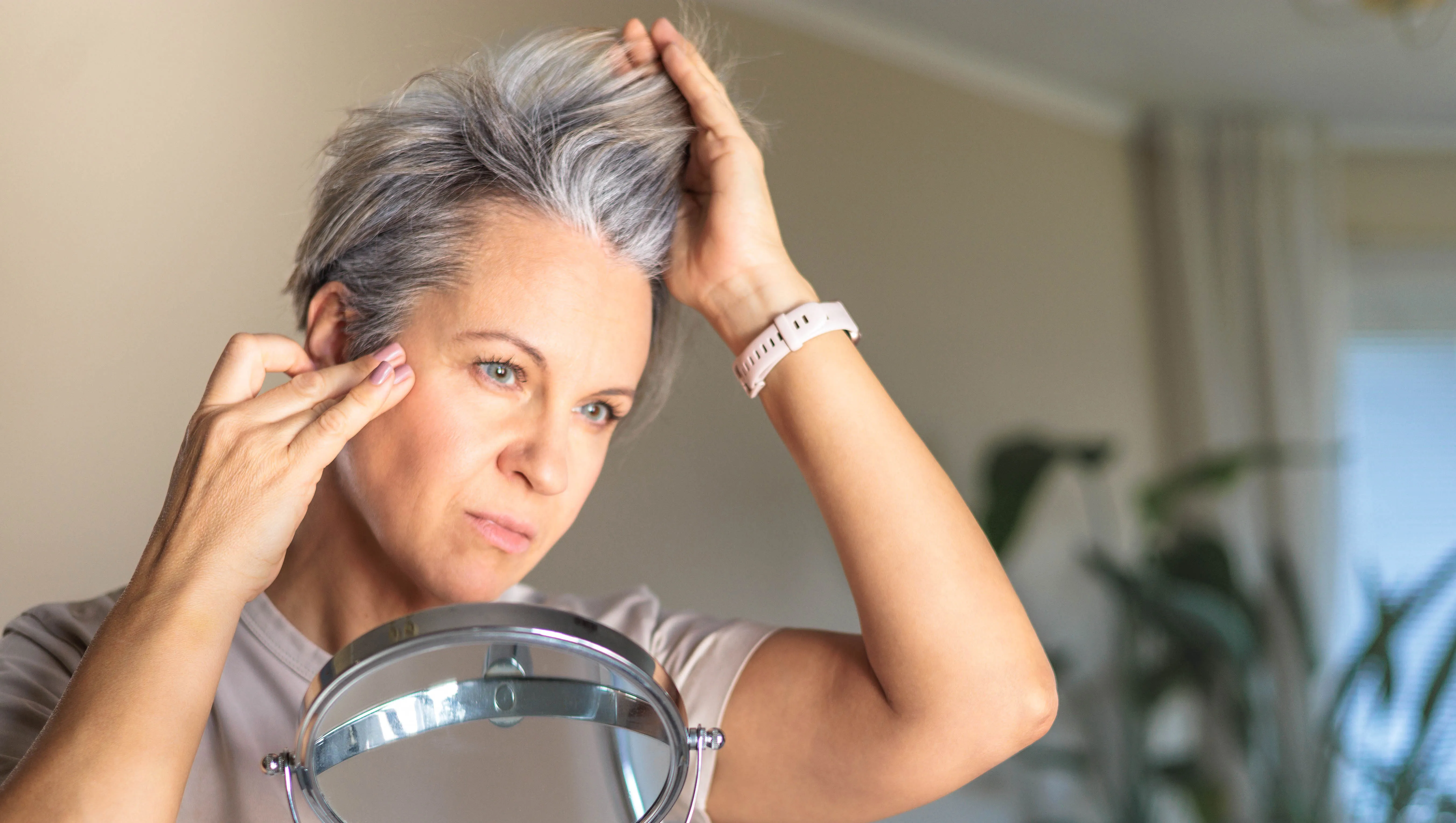 Gray-haired shocked middle-aged white woman looks at her gray hair and wrinkles in the mirror reflection. Concept aging and anti age.