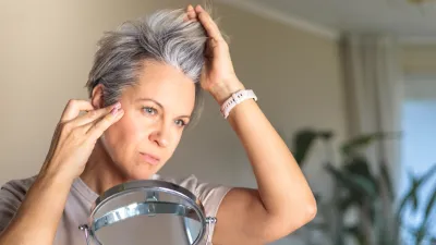 Gray-haired shocked middle-aged white woman looks at her gray hair and wrinkles in the mirror reflection. Concept aging and anti age.