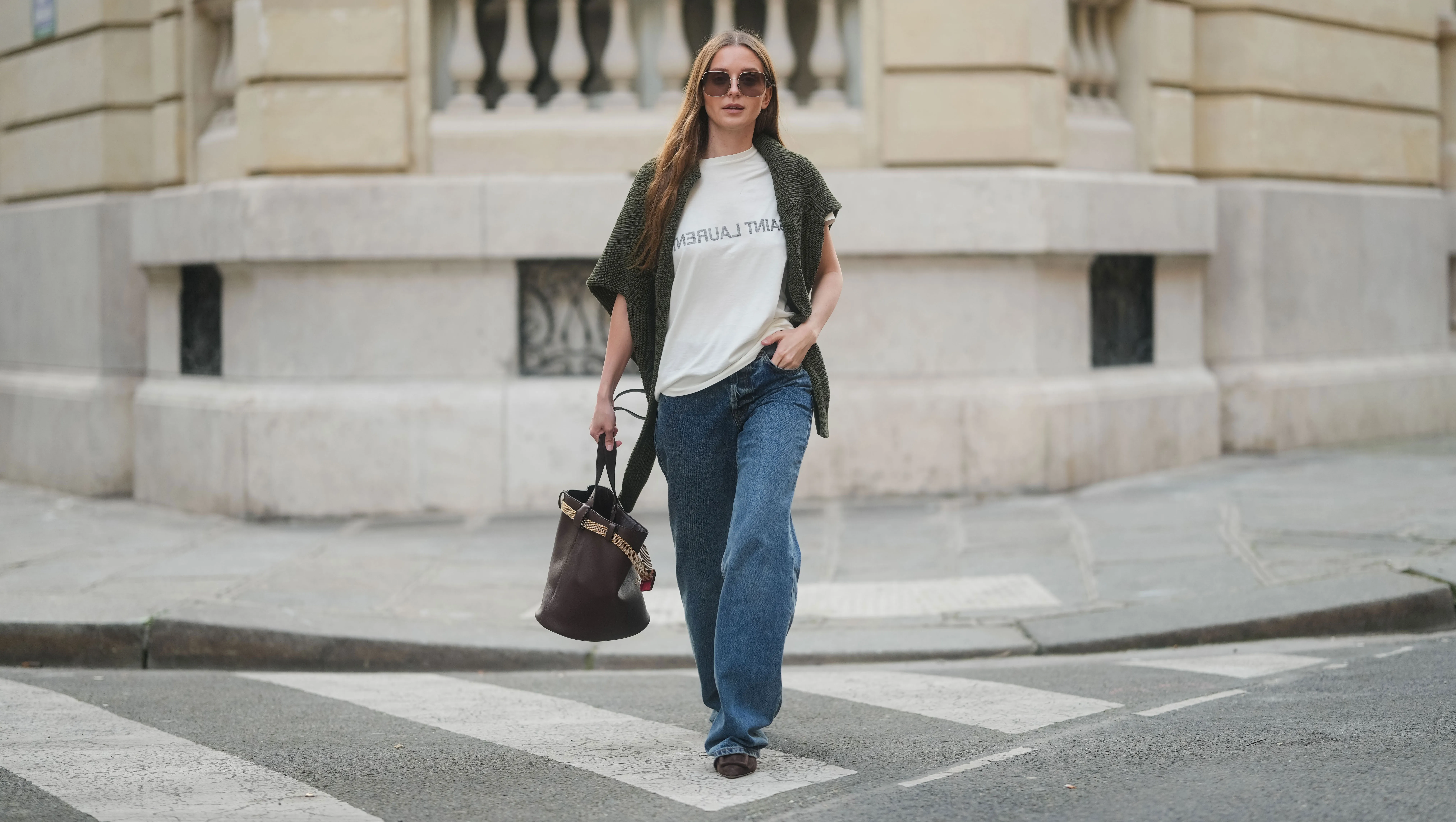 PARIS, FRANCE - MARCH 25: Diane Batoukina wears long straight brown hair with a center part, square Dior sunglasses, a dark green knit sweater by By Aylin Koenig draped over her shoulders, a white Saint Laurent T-shirt, wide-leg blue jeans by Anine Bing, brown suede pointed-toe flats by Manolo Blahnik, and carries a dark brown leather Prada bucket bag, during a street style fashion photo session on March 25, 2025 in Paris, France. (Photo by Edward Berthelot/Getty Images)