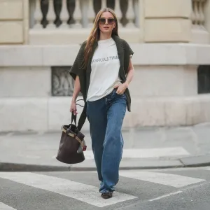 PARIS, FRANCE - MARCH 25: Diane Batoukina wears long straight brown hair with a center part, square Dior sunglasses, a dark green knit sweater by By Aylin Koenig draped over her shoulders, a white Saint Laurent T-shirt, wide-leg blue jeans by Anine Bing, brown suede pointed-toe flats by Manolo Blahnik, and carries a dark brown leather Prada bucket bag, during a street style fashion photo session on March 25, 2025 in Paris, France. (Photo by Edward Berthelot/Getty Images)