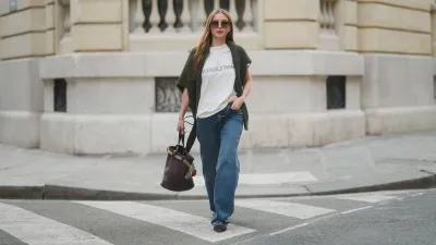 PARIS, FRANCE - MARCH 25: Diane Batoukina wears long straight brown hair with a center part, square Dior sunglasses, a dark green knit sweater by By Aylin Koenig draped over her shoulders, a white Saint Laurent T-shirt, wide-leg blue jeans by Anine Bing, brown suede pointed-toe flats by Manolo Blahnik, and carries a dark brown leather Prada bucket bag, during a street style fashion photo session on March 25, 2025 in Paris, France. (Photo by Edward Berthelot/Getty Images)