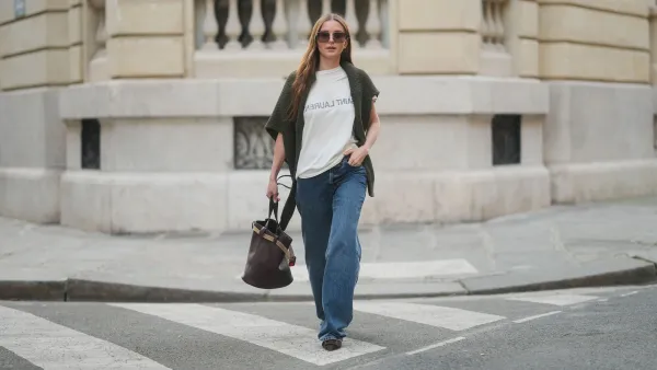 PARIS, FRANCE - MARCH 25: Diane Batoukina wears long straight brown hair with a center part, square Dior sunglasses, a dark green knit sweater by By Aylin Koenig draped over her shoulders, a white Saint Laurent T-shirt, wide-leg blue jeans by Anine Bing, brown suede pointed-toe flats by Manolo Blahnik, and carries a dark brown leather Prada bucket bag, during a street style fashion photo session on March 25, 2025 in Paris, France. (Photo by Edward Berthelot/Getty Images)
