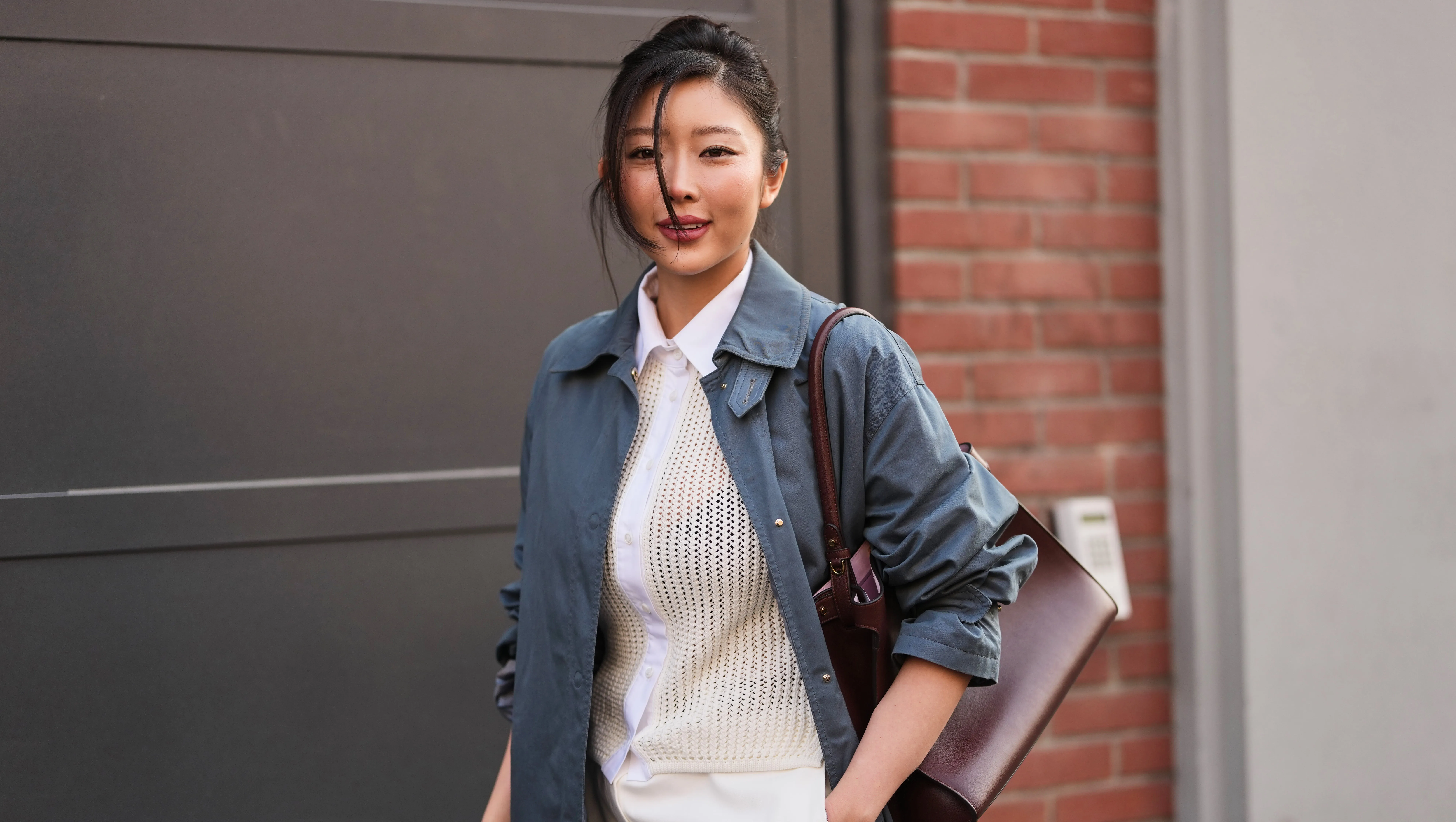 MILAN, ITALY - FEBRUARY 25: A guest wears black hair in a bun, a blue slate cotton trench coat, a white ivory cotton button down mesh / fishnet cardigan shirt, white ivory wide leg suit trousers, a brown burgundy leather shoulder bag, outside Fendi, during Milan Fashion Week - Womenswear Fall/Winter 2026/2027, on February 25, 2026 in Milan, Italy (Photo by Edward Berthelot/Getty Images)