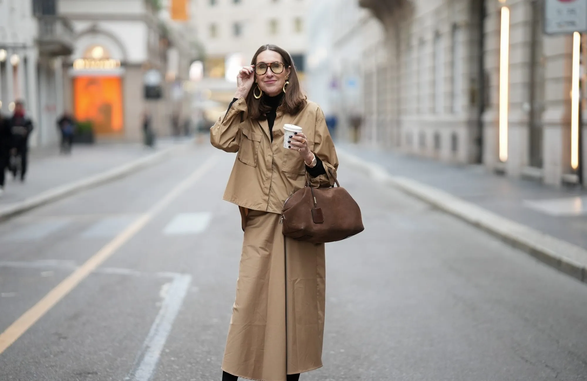 MILAN, ITALY - FEBRUARY 28: Annette Weber is seen wearing a camel colored button up overshirt with a relaxed silhouette and oversized flap chest pockets from Max Mara; a black fitted turtleneck from Max Mara; a matching camel colored midi skirt with a fluid fabric straight cut from Max Mara; a structured brown suede nubuk leather bag with top handles, a soft rounded shape and zip closure from Max Mara; sculptural oversized gold tone earrings with rounded elements from Max Mara; oversized eyeglasses with a tortoiseshell pattern frame from Mango; among her jewelry are chunky gold rings with diamond detailing; holding a coffee to go cup; her shoulder length brown hair is worn straight with a center part during the Milan Fashion Week - Womenswear Fall/Winter 2026/2027 on February 28, 2026 in Milan, Italy. (Photo by Moritz Scholz/Getty Images)