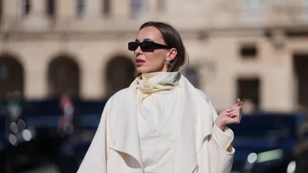 PARIS, FRANCE - MARCH 03: A guest wears black squared sunglasses, a gold logo and white pearls pendant earrings from Dior, a white wool long coat, black leather micro shorts, a white silk high neck blouse, outside Dior, during Paris Fashion Week - Womenswear Fall/Winter 2026/2027, on March 03, 2026 in Paris, France (Photo by Edward Berthelot/Getty Images)