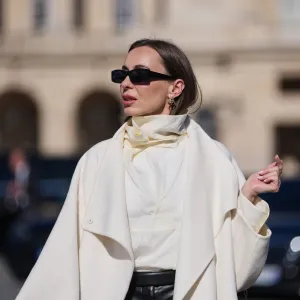 PARIS, FRANCE - MARCH 03: A guest wears black squared sunglasses, a gold logo and white pearls pendant earrings from Dior, a white wool long coat, black leather micro shorts, a white silk high neck blouse, outside Dior, during Paris Fashion Week - Womenswear Fall/Winter 2026/2027, on March 03, 2026 in Paris, France (Photo by Edward Berthelot/Getty Images)