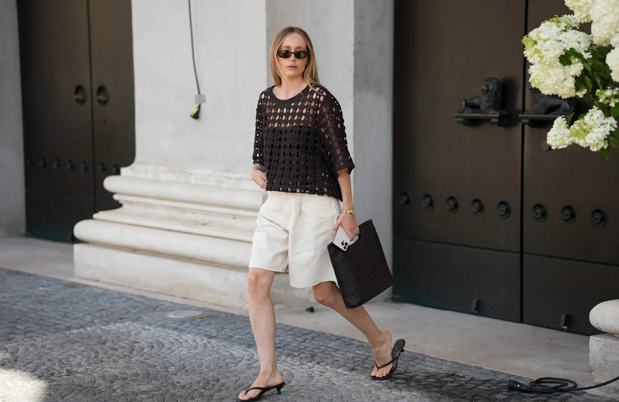 MUNICH, GERMANY - JULY 1: Anouk Delphine is seen wearing an H&M dark brown crochet t-shirt with three-quarter sleeves; a pair of beige Cos bermuda pants; a black squared weaved Cos hand bag; her blond hair is worn straight; black leather low heeled H&M thong sandals; a pair of oval Gucci sunglasses; a vintage gold Seiko watch; a Cartier light point gold necklace; Cartier bracelets; a gold Cartier brilliant-cut diamond ring paved with brilliant-cut diamonds; a Juste un Clou Cartier golden ring; she holds an iPhone in her hand on July 1, 2025 in Munich, Germany. (Photo by Moritz Scholz/Getty Images)