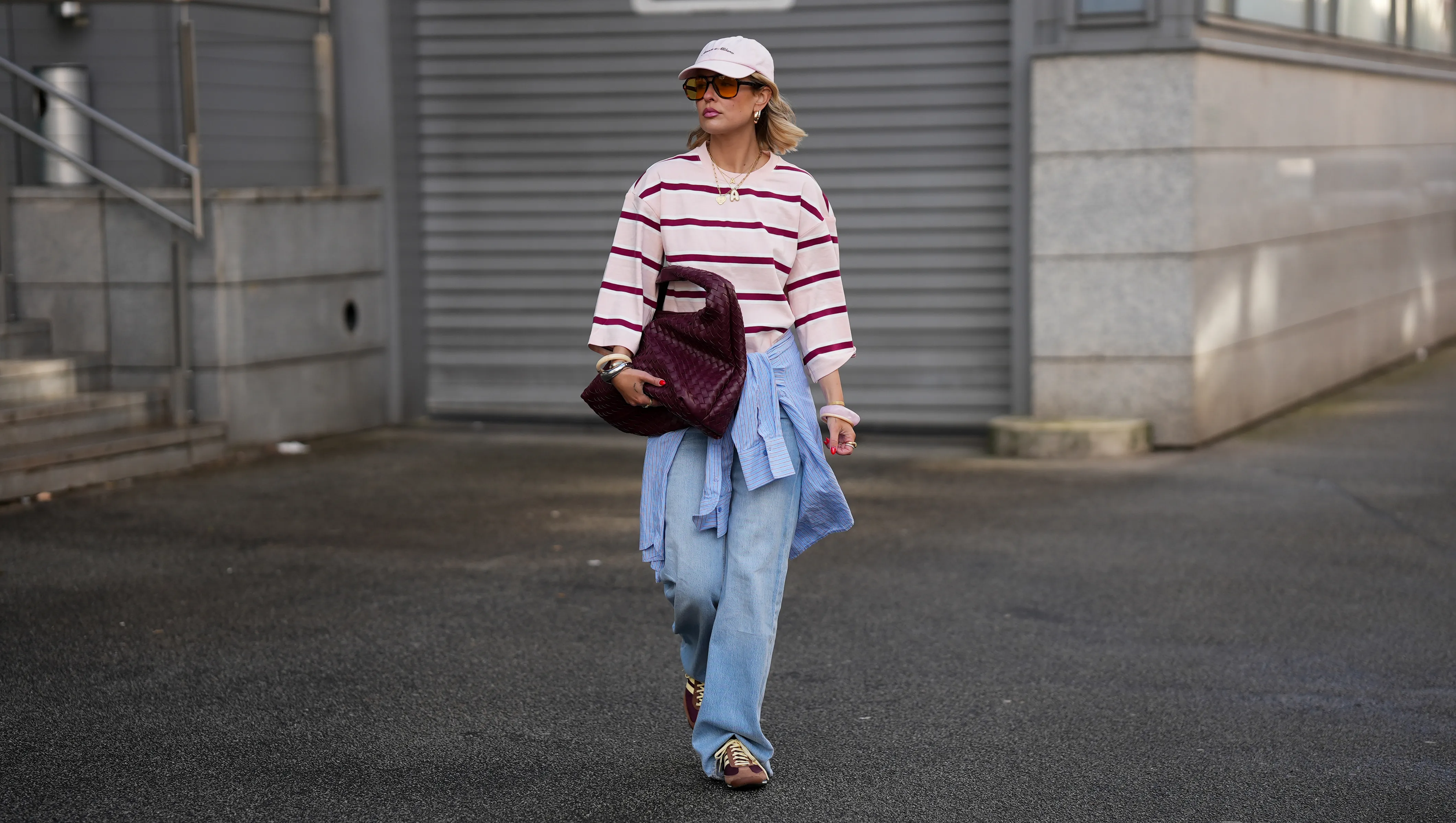 FRANKFURT AM MAIN, GERMANY - MARCH 26: Julia Kammerer is seen wearing an oversized pink and burgundy horizontal striped cotton long sleeve from H&M; a blue and white striped button down shirt tied around her waist from JJXX; light blue wide leg denim jeans with a relaxed fit from JJXX; burgundy and yellow textured leather lace up sneakers from Adidas; a large burgundy woven leather hobo bag from NA-KD; a pale pink baseball cap with the red embroidered text "Amore a Milano" from LeGer by Lena Gercke; her jewelry includes chunky gold hoop earrings, necklaces and gold tone stacked rings worn across multiple fingers from H&M, Pandora and My Jewellery; several silver and resin bangles from & Other Stories; orange tinted aviator sunglasses with a thin black frame from Bershka; her blonde hair is styled in loose waves underneath the cap on March 26, 2026 in Frankfurt am Main, Germany. (Photo by Moritz Scholz/Getty Images)