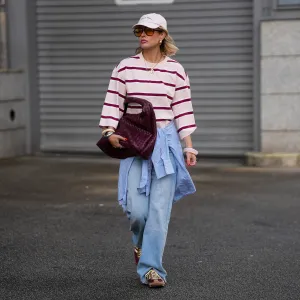 FRANKFURT AM MAIN, GERMANY - MARCH 26: Julia Kammerer is seen wearing an oversized pink and burgundy horizontal striped cotton long sleeve from H&M; a blue and white striped button down shirt tied around her waist from JJXX; light blue wide leg denim jeans with a relaxed fit from JJXX; burgundy and yellow textured leather lace up sneakers from Adidas; a large burgundy woven leather hobo bag from NA-KD; a pale pink baseball cap with the red embroidered text "Amore a Milano" from LeGer by Lena Gercke; her jewelry includes chunky gold hoop earrings, necklaces and gold tone stacked rings worn across multiple fingers from H&M, Pandora and My Jewellery; several silver and resin bangles from & Other Stories; orange tinted aviator sunglasses with a thin black frame from Bershka; her blonde hair is styled in loose waves underneath the cap on March 26, 2026 in Frankfurt am Main, Germany. (Photo by Moritz Scholz/Getty Images)