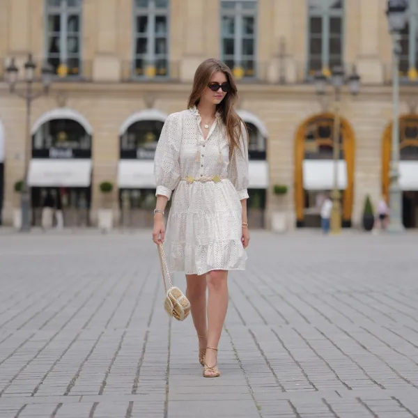 PARIS, FRANCE - JUNE 14: Segolene Hyppolite wears sunglasses, a white mini pleated and gathered summer dress with floral embroidery and long sleeves from Anne Fontaine, a rope belt from Anne Fontaine, a raffia bag from Vanessa Bruno, golden sandals / shoes from Sezane, during a street style fashion photo session, on June 14, 2024 in Paris, France. (Photo by Edward Berthelot/Getty Images)