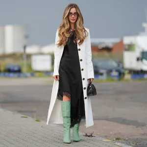 COPENHAGEN, DENMARK - AUGUST 07: Rosa Crespo wears a long white coat with large brown buttons, layered over a black dress featuring lace trim at the hem. The outfit is accessorized with a small black textured handbag. Light green knee-high boots complete the ensemble. Hair is styled in loose waves, and dark-framed glasses are worn, outside Rotate, during Copenhagen Fashion Week, on August 07, 2025 in Copenhagen, Denmark (Photo by Edward Berthelot/Getty Images)