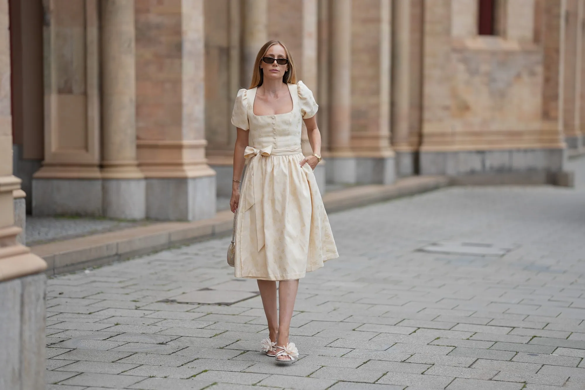 MUNICH, GERMANY - SEPTEMBER 12: Anouk Delphine is seen wearing a cream-colored midi dress with subtle floral jacquard, puff sleeves, a square neckleine and a waist bow from Limberry; open-toe heeled sandals in a soft ivory shade, detailed with oversized floral appliqués from Zara; a small structured handbag in the shape of a heart covered in shimmering metallic texture from Weat; a golden watch with a metallic wristband from Vintage Seiko; narrow dark sunglasses with brown tinted lenses from Gucci; her blonde hair is worn loose on September 12, 2025 in Munich, Germany. (Photo by Moritz Scholz/Getty Images)