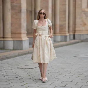 MUNICH, GERMANY - SEPTEMBER 12: Anouk Delphine is seen wearing a cream-colored midi dress with subtle floral jacquard, puff sleeves, a square neckleine and a waist bow from Limberry; open-toe heeled sandals in a soft ivory shade, detailed with oversized floral appliqués from Zara; a small structured handbag in the shape of a heart covered in shimmering metallic texture from Weat; a golden watch with a metallic wristband from Vintage Seiko; narrow dark sunglasses with brown tinted lenses from Gucci; her blonde hair is worn loose on September 12, 2025 in Munich, Germany. (Photo by Moritz Scholz/Getty Images)